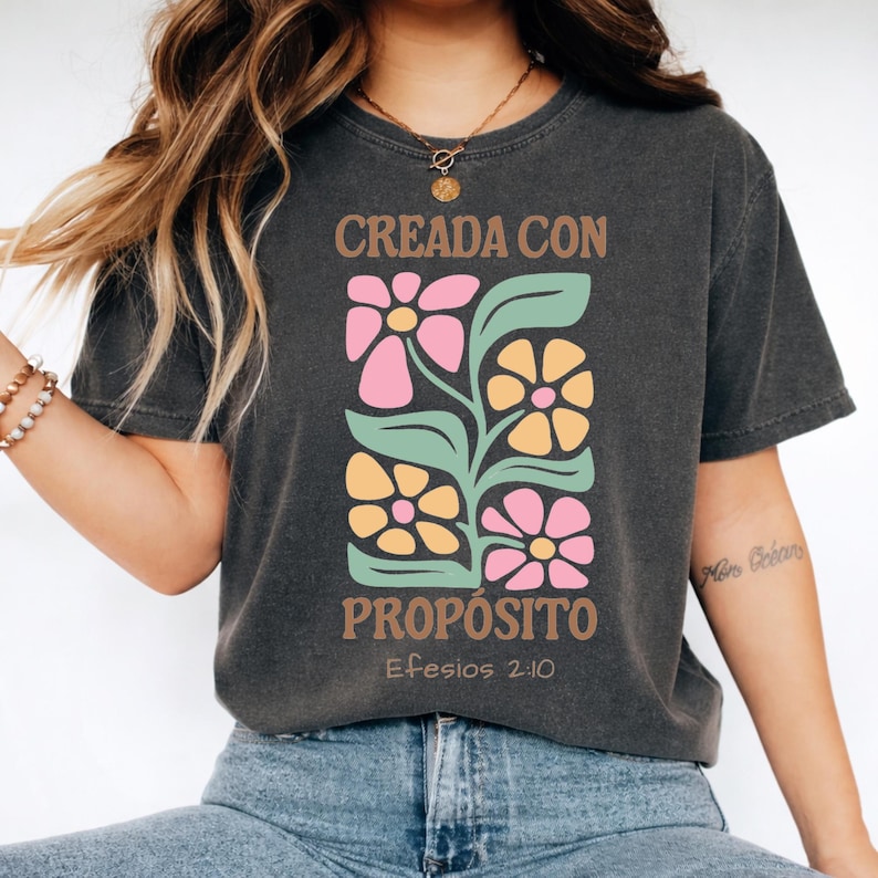 May include: Dark gray t-shirt with a floral design in pink, yellow, and green. The shirt features the text "CREADA CON PROP&Oacute;SITO" and "Efesios 2:10" in brown. The design is centered on the front of the shirt.