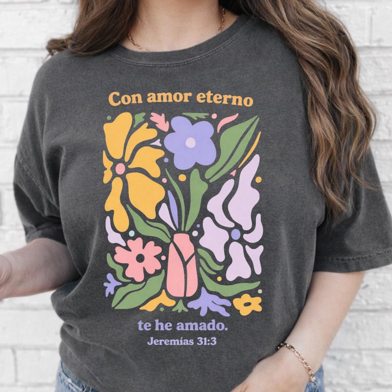 May include: Dark gray t-shirt featuring a colorful floral design with the text "Con amor eterno te he amado. Jerem&iacute;as 31:3". The design includes yellow, purple, pink, and green flowers in a vase.