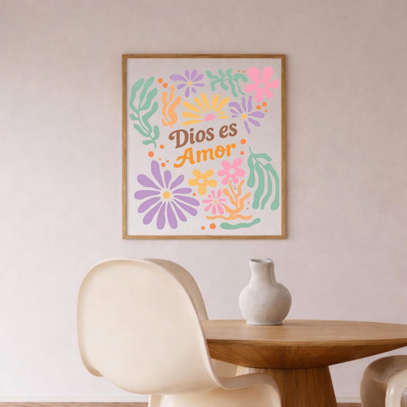 May include: A framed art print with the text "Dios es Amor" surrounded by colorful floral illustrations in shades of purple, pink, yellow, and green. The print is displayed on a white wall above a wooden table and a white vase.