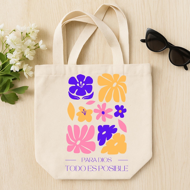May include: Cream-colored tote bag with a floral design in purple, yellow, and pink. The bag features the text "PARA DIOS TODO ES POSIBLE" in purple. Black sunglasses rest on a light wooden surface.