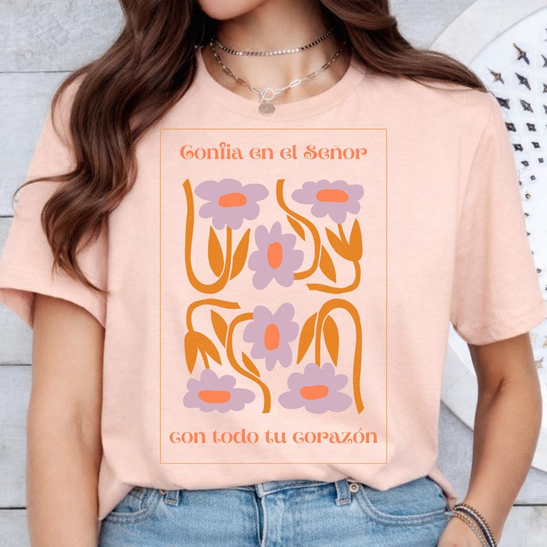 May include: A peach-colored t-shirt featuring a floral design with the text "Conf&iacute;a en el Se&ntilde;or" and "Con todo tu coraz&oacute;n" in a rectangular frame. The design includes purple flowers with orange centers and stems.