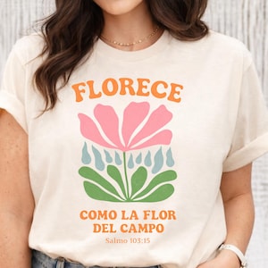 May include: Cream-colored t-shirt with the word "FLORECE" in orange above a pink flower with blue raindrops and green leaves. The text "COMO LA FLOR DEL CAMPO" and "Salmo 103:15" are printed below the flower.