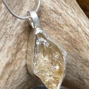 May include: A pendant necklace featuring a raw, translucent yellow crystal encased in a textured silver setting. The crystal has a jagged, irregular shape with internal facets that catch the light. It hangs from a thin, flexible silver chain.