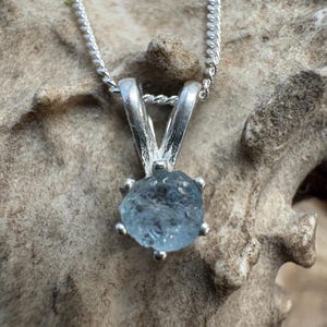 May include: A silver necklace featuring a raw, light blue aquamarine gemstone pendant. The pendant is held in a silver setting with six prongs. The chain is a delicate silver cable chain. The background is a textured, neutral surface.