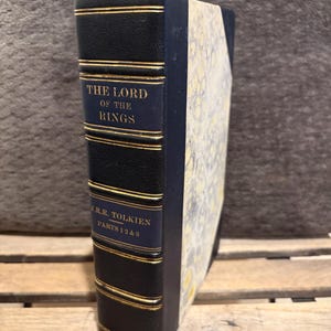 May include: A vintage hardcover edition of "The Lord of the Rings" by J.R.R. Tolkien. The book features a dark blue leather spine with gold lettering and trim. The cover is a light colour with a floral pattern. The book is from 1968.