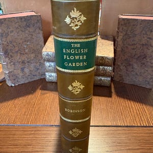 May include: A tall, antique book titled "The English Flower Garden" by Robinson. The book has a brown leather cover with gold floral accents and a green band with gold lettering. The book is standing upright on a wooden surface.