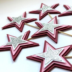 May include: Several layered star-shaped cupcake toppers. Each star has a dark red outer layer, a pink middle layer, a white inner layer, and a silver glitter star in the center. Wooden toothpicks are attached to the bottom.