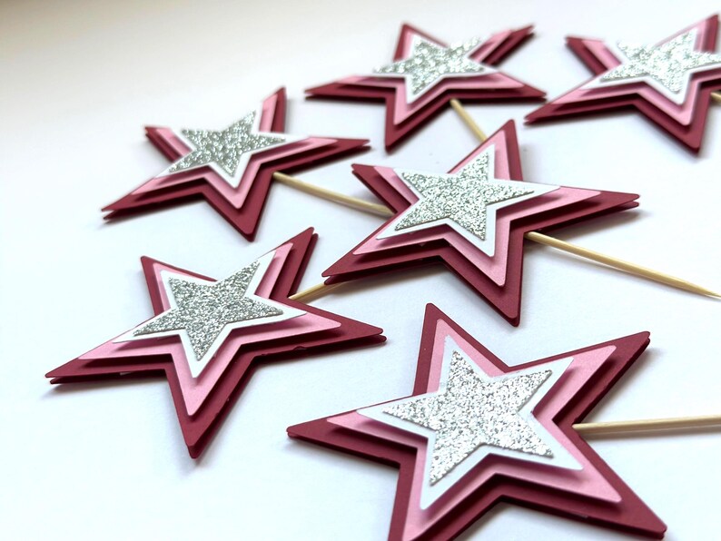 May include: Several layered star-shaped cupcake toppers. Each star has a dark red outer layer, a pink middle layer, a white inner layer, and a silver glitter star in the center. Wooden toothpicks are attached to the bottom.