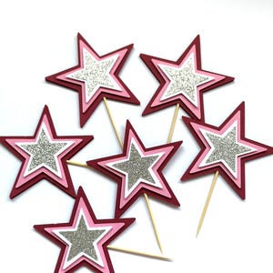 May include: A collection of star-shaped cupcake toppers. Each star has a layered design with a glittery silver center, surrounded by white, pink, and burgundy layers. The toppers are attached to wooden sticks.