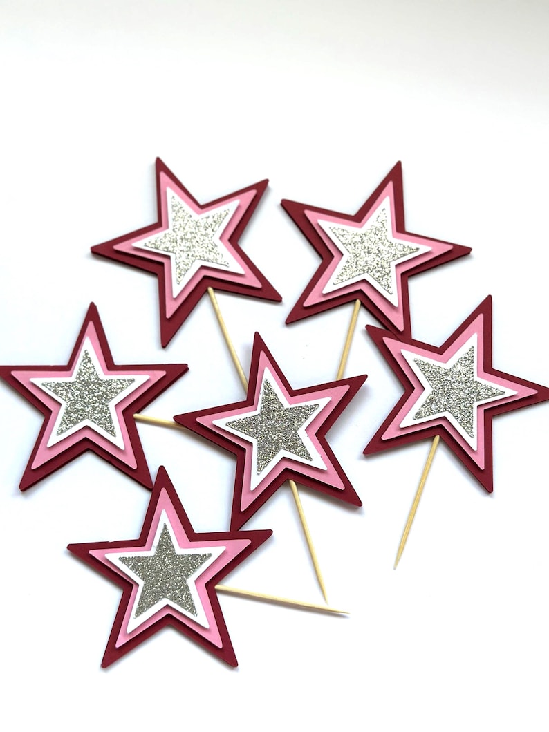 May include: A collection of star-shaped cupcake toppers. Each star has a layered design with a glittery silver center, surrounded by white, pink, and burgundy layers. The toppers are attached to wooden sticks.