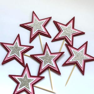 May include: Six star-shaped cake toppers. Each star has a layered design with a dark red outer layer, a pink middle layer, a white inner layer, and a silver glitter center. The toppers are attached to wooden sticks.