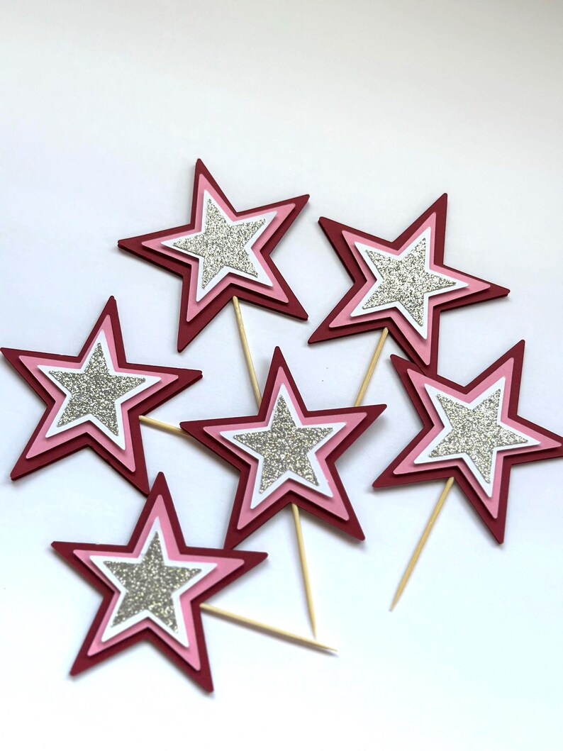 May include: Six star-shaped cake toppers. Each star has a layered design with a dark red outer layer, a pink middle layer, a white inner layer, and a silver glitter center. The toppers are attached to wooden sticks.