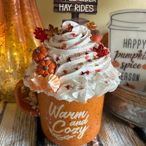 May include: An orange mug with the words "Warm and Cozy" in white script, topped with whipped cream, pumpkins, and fall leaves. A sign above reads "Pumpkins, Apple Cider, Hay Rides".