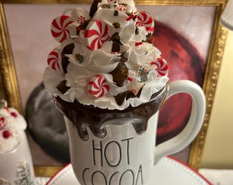 May include: A white ceramic mug with the text HOT COCOA in black lettering, topped with faux whipped cream, chocolate drizzle, red and white peppermint candies, and two candy canes. The mug features a pedestal base and a curved handle.