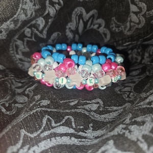 May include: A stack of colorful beaded bracelets. The bracelets feature pink, light blue, and white beads, with clear star-shaped accents. The word "MIKU" is spelled out with letter beads. The bracelets are stacked on top of each other.