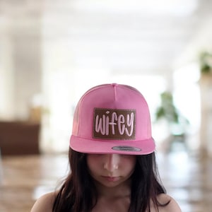 May include: A pink baseball cap with a brown patch displaying the word "wifey" in white script. The cap features a mesh back and a curved peak. The hat is a solid pink colour.