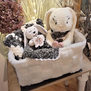 May include: A white crochet bunny with a black and white scarf and a smaller bunny with a pink flower, nestled in a white faux fur basket with a black and white blanket. The basket is on a wooden table.