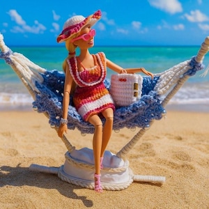 以下が含まれることがあります： 砂浜の青と白の編み物ハンモックに座っている、かぎ針編みのドレス、帽子、ピンクのサングラスを身に着けた人形。 人形は白いかぎ針編みのバッグを持っています。 背景には青い海と空があります。