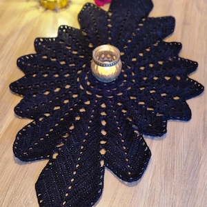 May include: A navy blue, handmade crochet table runner in a floral design. The runner has a central circular area with a candle holder and a scalloped edge. The table runner is on a wooden surface.