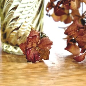 Peut inclure: Gros plan sur une fleur d'hortensia séchée, brun rougeâtre, avec des gouttelettes d'eau, posée sur une surface en bois. Un vase décoratif en verre avec un motif tourbillonnant est en arrière-plan, avec d'autres fleurs séchées.
