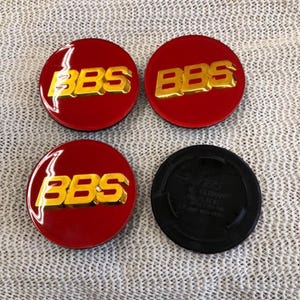 May include: Four red and gold BBS wheel center caps. The caps have a glossy finish with gold lettering. One black cap is also included. The caps are likely for a car or vehicle.