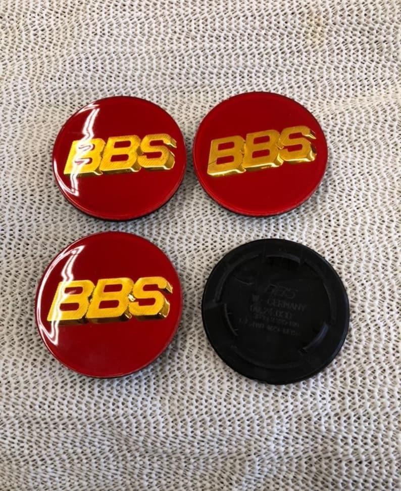 May include: Four red and gold BBS wheel center caps. The caps have a glossy finish with gold lettering. One black cap is also included. The caps are likely for a car or vehicle.