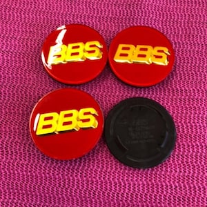 May include: Four red and gold BBS wheel center caps. Three are circular with the BBS logo in gold. One black cap is also included. The caps are on a pink textured surface.