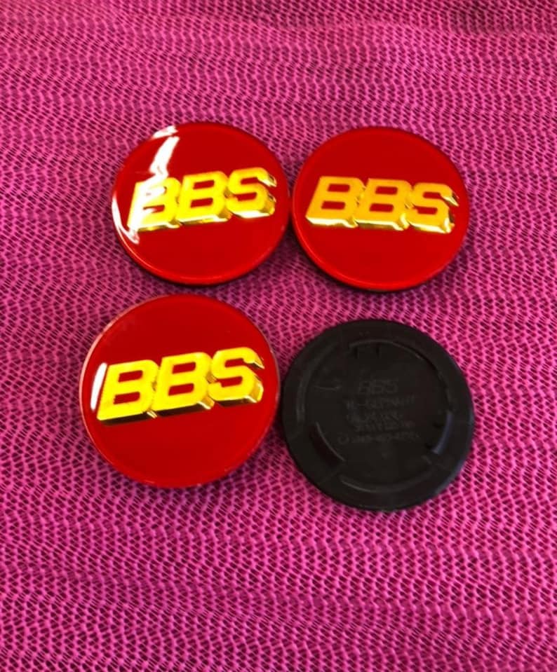 May include: Four red and gold BBS wheel center caps. Three are circular with the BBS logo in gold. One black cap is also included. The caps are on a pink textured surface.
