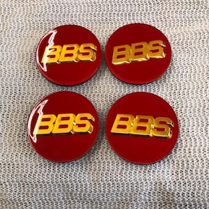 Puede incluir: Cuatro emblemas circulares rojos con las letras doradas "BBS" en el centro. Los emblemas son brillantes y parecen estar hechos de plástico duro o metal. El fondo es una superficie texturizada gris claro.