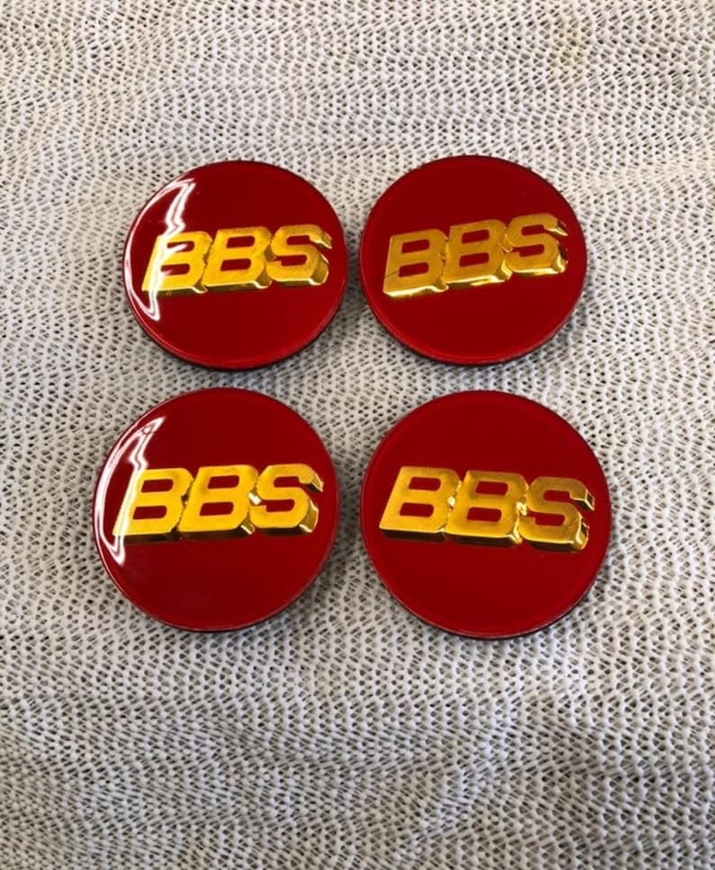 May include: Four red circular emblems with the gold letters "BBS" in the center. The emblems are glossy and appear to be made of a hard plastic or metal. The background is a light gray, textured surface.