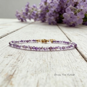 May include: Delicate amethyst bead bracelet with a gold clasp. The bracelet is strung with small, faceted purple beads. The background features a blurred arrangement of purple flowers and a white wooden surface. The text "Onyx The Finest" is visible.