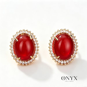 May include: A pair of gold-toned stud earrings. Each earring features a large, oval, deep red gemstone surrounded by a halo of small, clear crystals. The earrings are set against a white background, with the brand name "ONYX THE FINEST" visible.
