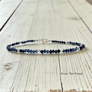 May include: A delicate anklet featuring small, faceted, dark blue beads interspersed with silver-colored accents. The anklet is secured with a silver-toned clasp. The background is a light-colored wooden surface. The text "Onyx The Finest" is visible.