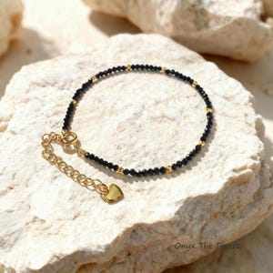 May include: Black beaded bracelet with gold accents and a heart charm. The bracelet is made of small, round black beads interspersed with gold beads. The clasp and heart charm are gold-colored. The bracelet is displayed on a white, textured surface.
