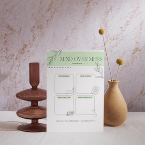 May include: A white notepad with the text "MIND OVER MESS" and four boxes for notes. The notepad is decorated with green leaves and is next to a brown vase with yellow flowers and a brown glass decorative object.