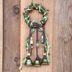 May include: A handcrafted wall hanging featuring a circular grapevine wreath adorned with faux green leaves and brown twine. Three bronze-coloured bells hang below, accompanied by decorative charms and small glass bottles. The piece is mounted on a wooden background.