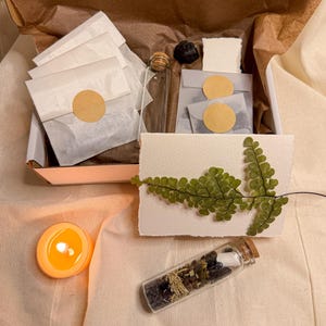 May include: An open box containing a collection of items: small glass bottles with corks, white paper packets with circular stickers, and a small lit orange candle. A sprig of green leaves decorates a piece of white paper.