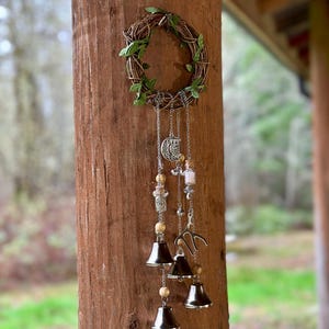 Puede incluir: Un carillón rústico con una pequeña corona de ramitas y hojas verdes. Cadenas plateadas cuelgan, adornadas con amuletos, cuentas de madera y campanas plateadas. El carillón está sujeto a un poste de madera.