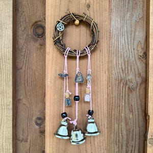 Puede incluir: Una corona circular de vid con detalles de cuerda rosa. La corona presenta colgantes, pequeñas botellas de vidrio y campanas plateadas. Los colgantes incluyen un pentagrama, un murciélago, un ojo y una llave. Las campanas son plateadas y tienen un acabado brillante.