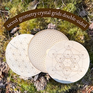 May include: Three double-sided wooden crystal grids with geometric designs. The grids feature the Flower of Life, a radial pattern, and Metatron's Cube. The grids are light-colored and set on a bed of green moss.