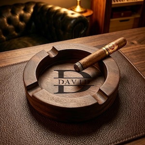 May include: A wooden ashtray with the name "DAVID" engraved in black. A lit cigar rests on the ashtray, which is placed on a brown leather surface. The ashtray has four grooves for holding cigars.