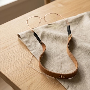 May include: A pair of rose gold metal spectacles attached to a tan leather strap. The strap features black leather ends secured with metallic rivets and the letters JSN embossed in gold on the bottom centre of the leather band.