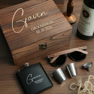 Könnte beinhalten: Eine Holzkiste, eine schwarze Flasche, Sonnenbrillen und Schnapsgläser sind auf einer Holzoberfläche angeordnet. Die Kiste und die Flasche sind mit dem Namen "Gavin" und dem Text "Groomsman 05.30.2026" graviert. Die Sonnenbrille hat Holzgestelle.
