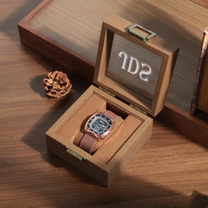 Può includere: Un orologio in oro rosa con quadrante nero e cinturino in pelle marrone è contenuto in una scatola di legno aperta con coperchio in vetro. L'interno della scatola è marrone chiaro. Una pigna è a sinistra.