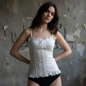 Puede incluir: Un camisón de encaje blanco con finas tiras de espagueti, un escote con volantes y una fila de pequeños botones en la parte delantera. El camisón tiene una silueta ajustada y un dobladillo festoneado. La modelo lleva ropa interior negra.