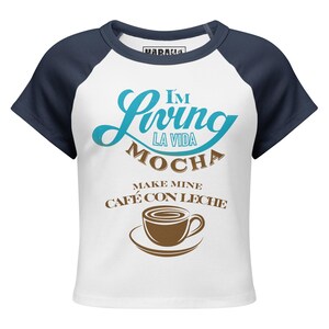 May include: White and navy blue short-sleeved t-shirt featuring the text "I'm Living La Vida Mocha, Make Mine Café Con Leche" and a coffee cup graphic. The shirt has a crew neck and raglan sleeves.