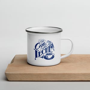 Op de afbeelding: Witte emaille mok met een zilveren rand, met de tekst "Love is Cafe con Leche" in blauw schrift, samen met een blauwe illustratie van een vogel en een koffiekopje. De mok staat op een houten oppervlak.