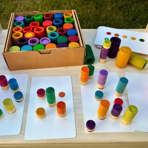 Könnte beinhalten: Ein Holzspielzeug-Set mit farbenfrohen Zylindern und Scheiben in einem Karton, mit zusätzlichen Teilen und passenden Karten. Die Zylinder sind in verschiedenen Farben lackiert. Lernspielzeug für Kinder.