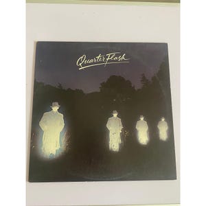 May include: Album cover for Quarterflash, featuring a dark, nighttime scene. Silhouetted figures in coats and hats stand in a row, illuminated by a soft glow. The band's name is written in cursive across the top.
