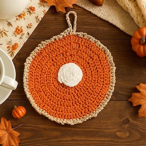 May include: A handmade, round orange crochet trivet with a white center, perfect for fall decor. The trivet has a tan border and a loop for hanging. Decorative pumpkins, leaves, and a teapot are also visible.
